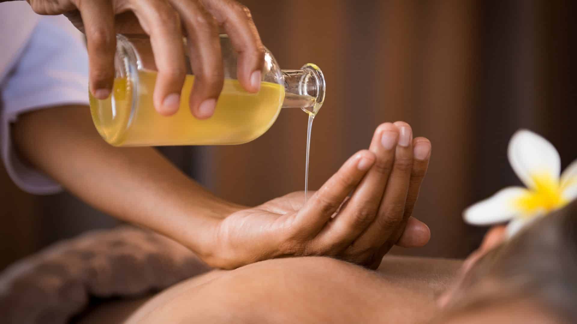 hand pouring massage oil into another hand over the back of a massage patient who has a white and yellow flower behind their ear