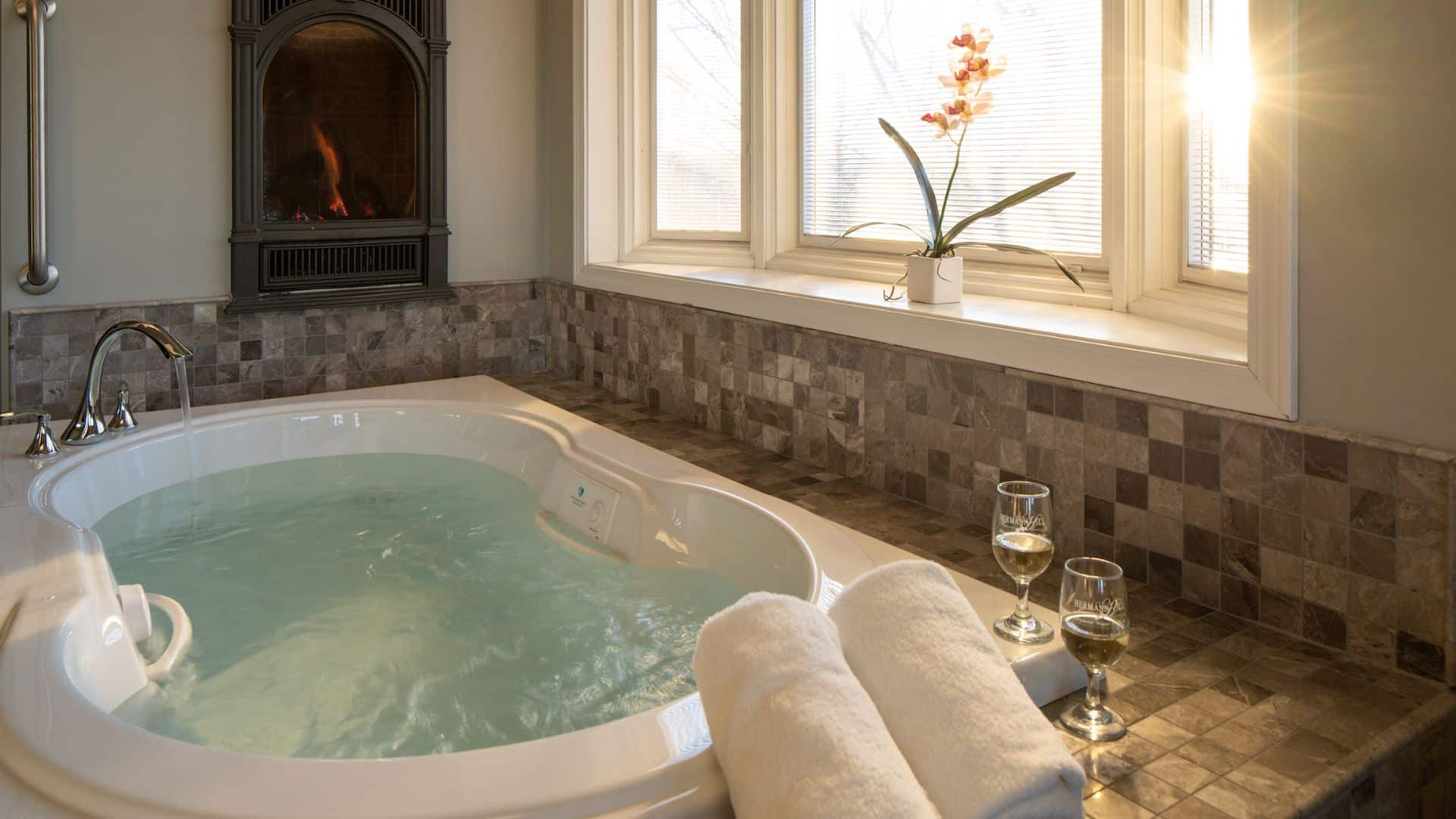 Jetted tub surrounded by tile, a bay window, a fireplace, and two wine glasses.