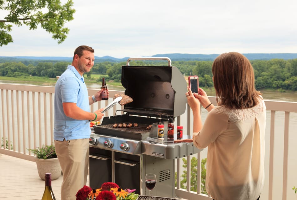 Man and woman on large deck smiling and grilling food with a river and trees in the background