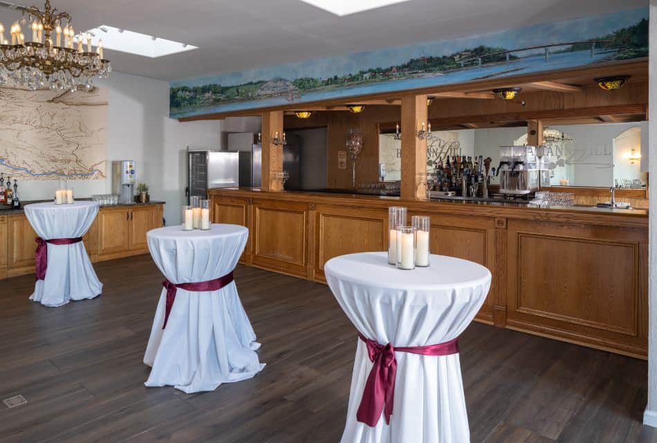 Large room with a wooden bar and three cocktail tables draped with white tablecloths and wrapped with burgundy ribbons tied in a bow