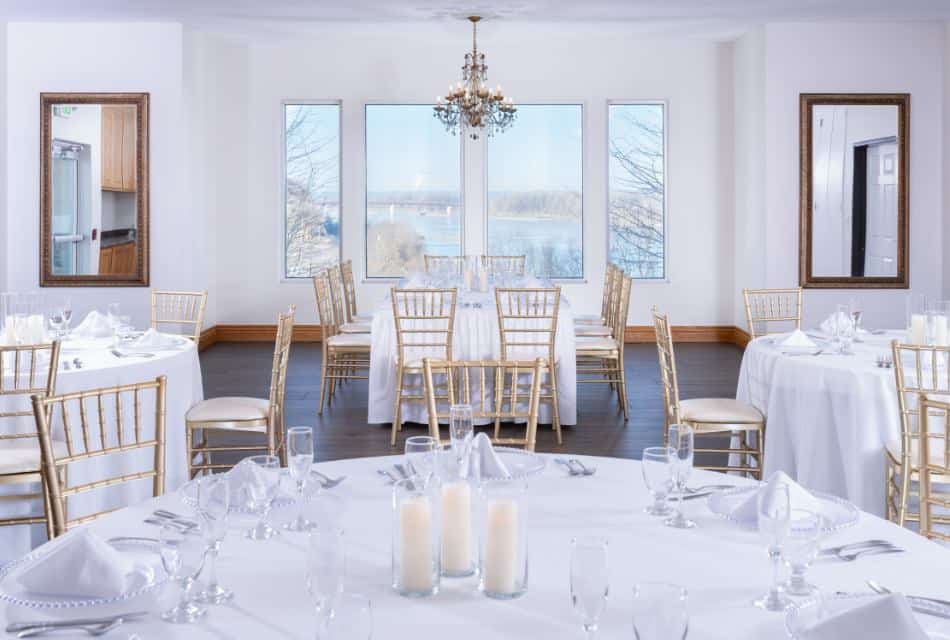 Large banquet hall with gold chairs, tables with white tablecloths, full placesettings, and windows showing the river