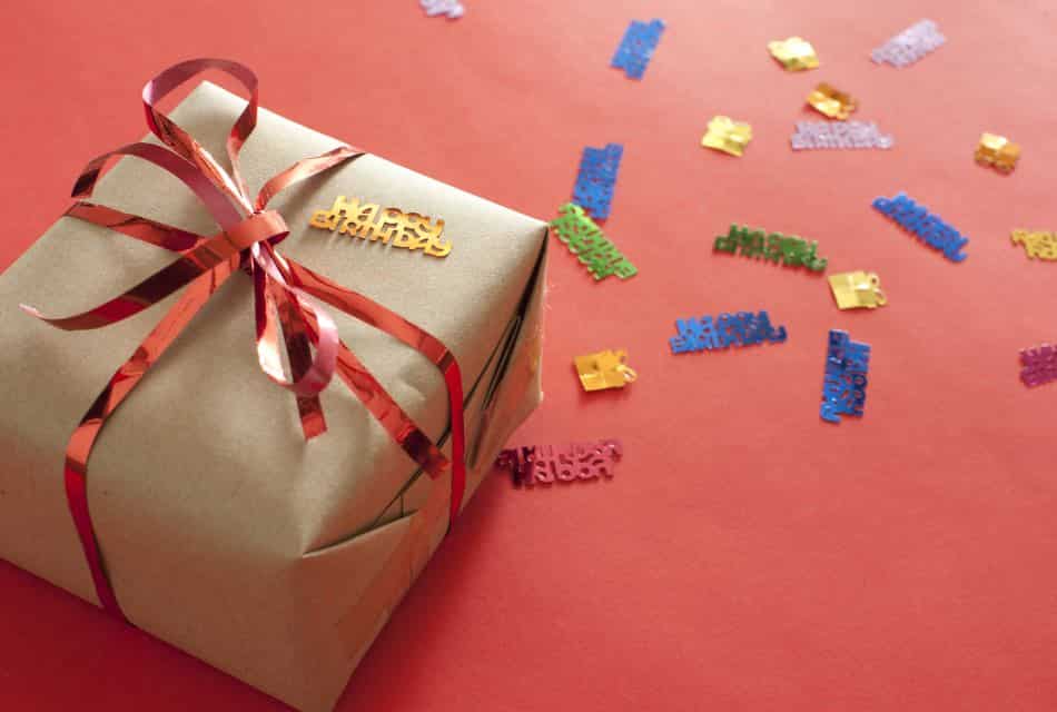 Wrapped package in kraft paper with red ribbon on red tablecloth with Happy Birthday confetti on the table