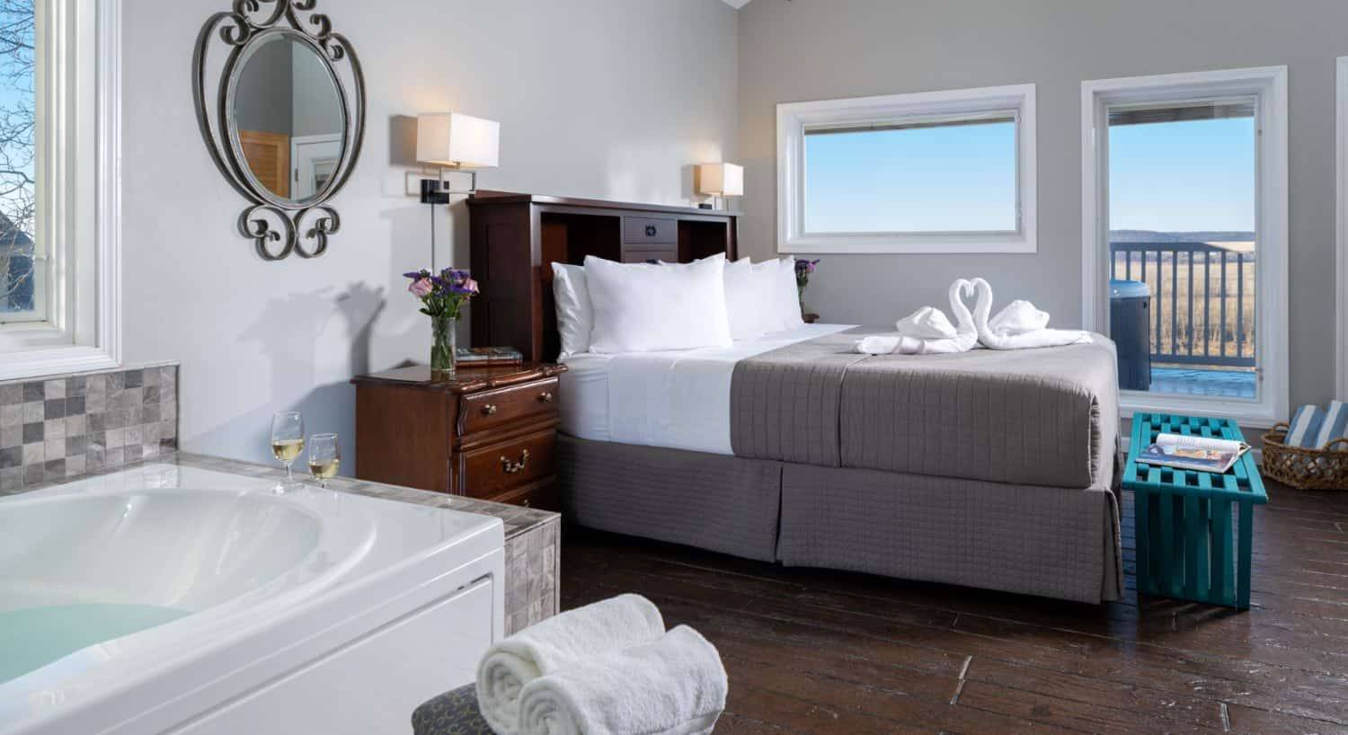 Large bedroom suite with light gray walls, wooden flooring, wooden headboard, white bedding, gray comforter, and jetted tub