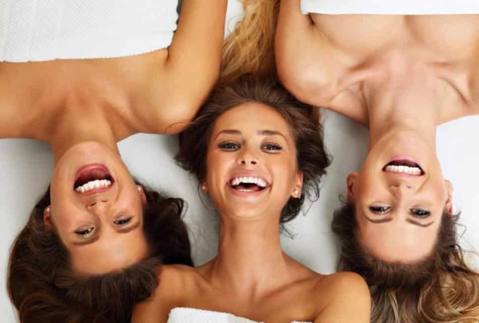 Three women wearing white towels laying on white bedding allm smiling and looking at the camera