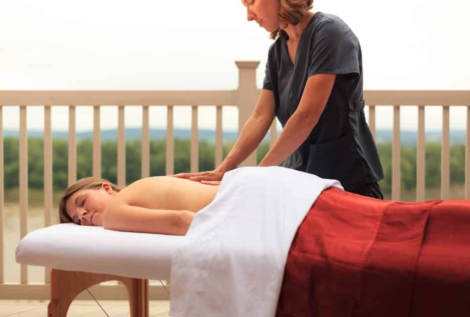 Woman in gray shirt giving a massage to a woman laying down on massage table covered by a white and red blanket